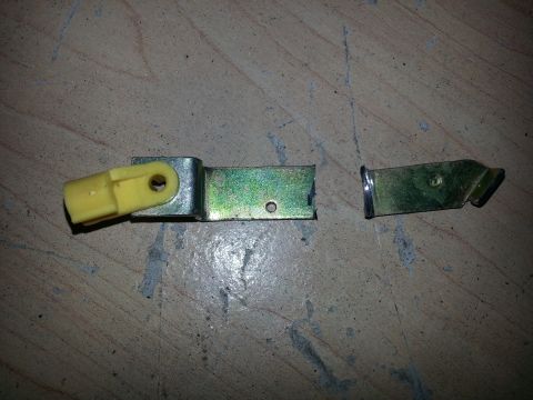 1994 Ford Bronco - A broken tailgate latch mechanism, consisting of two metal pieces and a yellow plastic component, lies on a wooden surface.