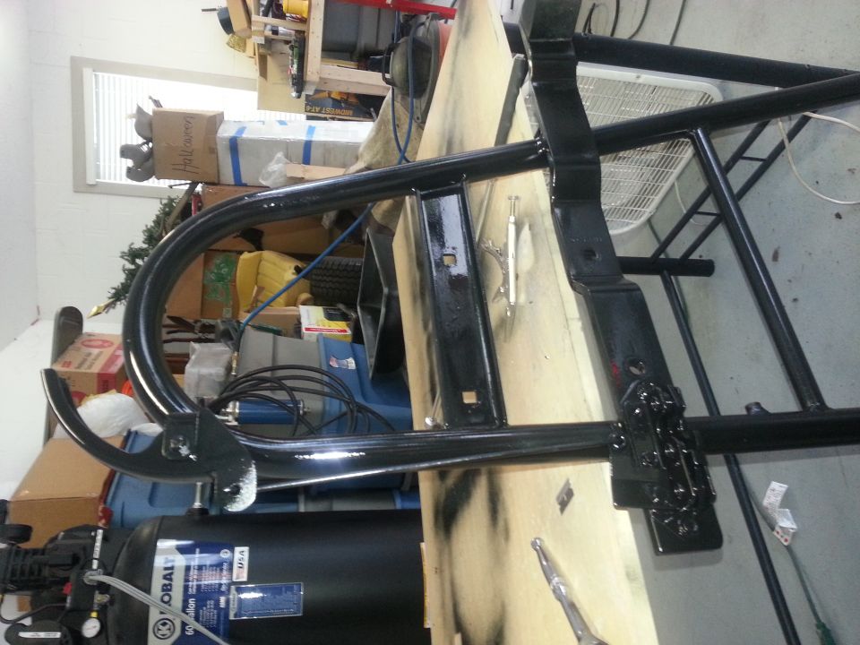Black metal frame part, possibly a roll cage or bumper, on a workbench with an air compressor in the background