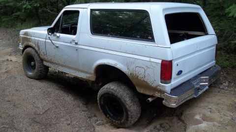 1990 Ford Bronco - A white Ford Bronco navigates a muddy off-road trail, its tires kicking up dirt.