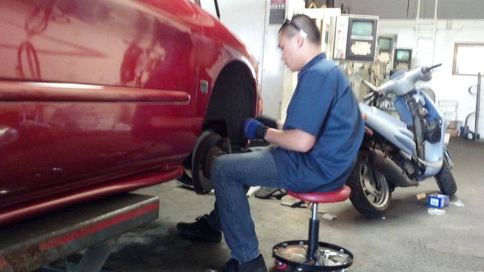 Red car side view, mechanic working on wheel well. Scooter in background