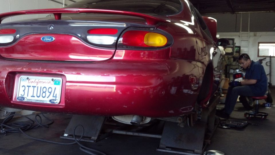 Rear of a red 1996 Ford Taurus on lifts in a garage with a mechanic working nearby