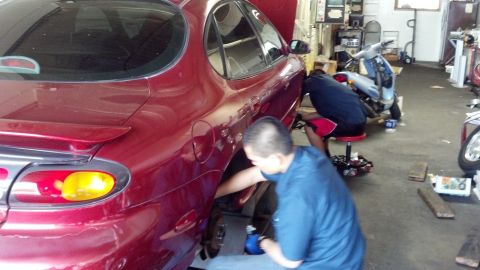 1996 Ford Taurus - Two mechanics are working on a red sedan in a garage, with a scooter visible in the background.