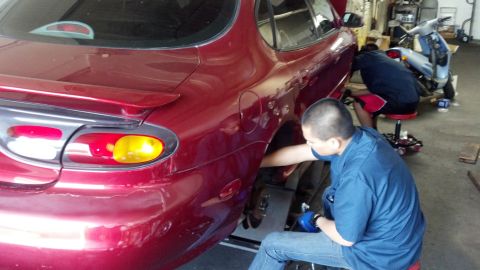 1996 Ford Taurus - Mechanics work on the rear wheel of a red sedan, possibly a 1995 model, in a garage with a scooter nearby.