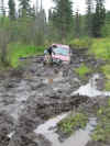 1979 Ford Bronco - An off-road vehicle navigates a challenging muddy trail, with dense foliage surrounding the path.