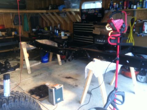 1979 Ford Bronco - The frame of a vehicle is elevated on sawhorses in a workshop setting, surrounded by various tools and supplies.
