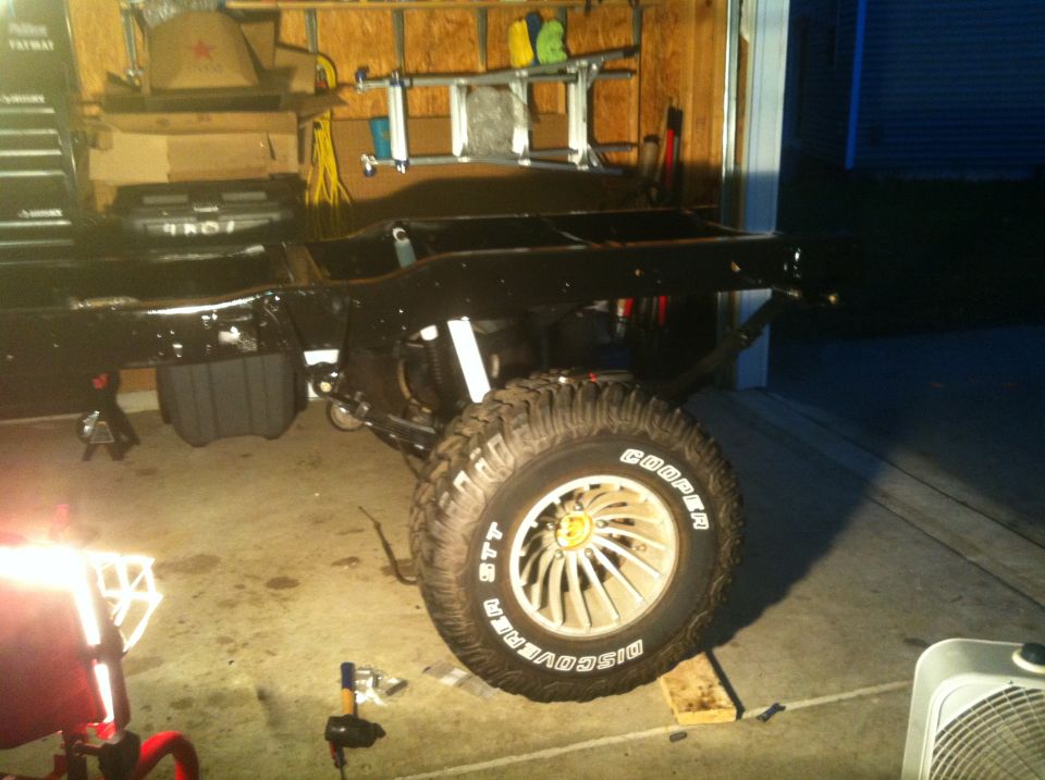 Black vehicle frame and suspension with large off-road tire on a jack stand in a garage