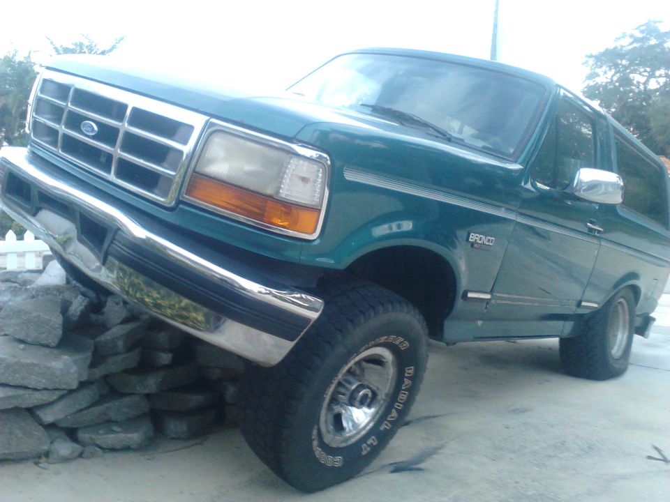 Green Ford Bronco SUV with front tire on a rock pile, low 3/4 front view showing chrome grille and white-letter tires