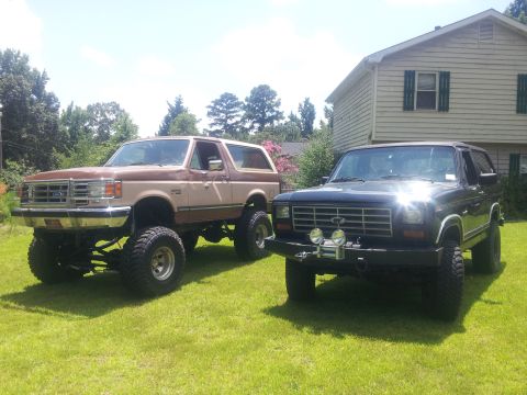 1986 Ford Bronco - Two lifted Ford Broncos sit on a grassy lawn on a sunny day, with a house in the background.