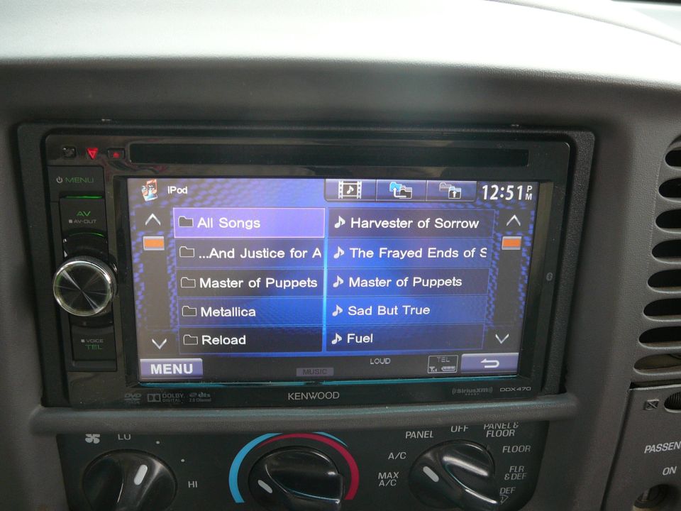 Car stereo screen displaying iPod menu with song titles like 'All Songs' and 'Master of Puppets'