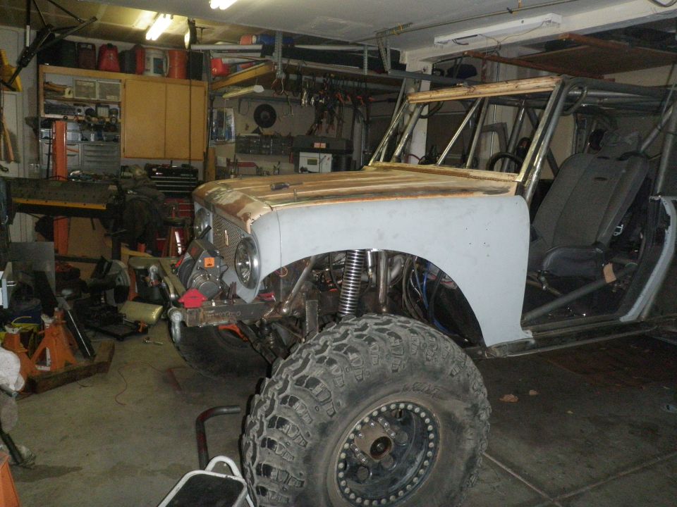 Off-road vehicle body in primer, large tire, roll cage, and racing seat visible in a garage