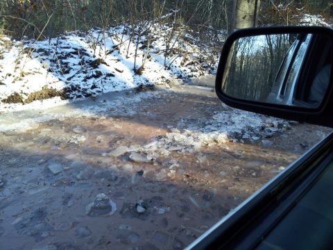 2003 Nissan Frontier - The side view mirror reflects trees as the vehicle navigates a muddy and icy road, suggesting off-road conditions.