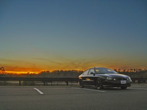 1997 Ford Taurus - A black sedan is parked in a lot during sunset, with a highway barrier in the background.