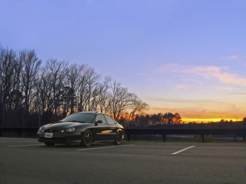 1997 Ford Taurus - A black 1997 Ford Taurus is parked in a lot as the sun sets behind a line of trees.