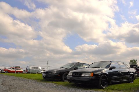 1997 Ford Taurus - Two black sedans are parked on a gravel lot under a cloudy sky, with a vintage station wagon and an Airstream trailer in the background.