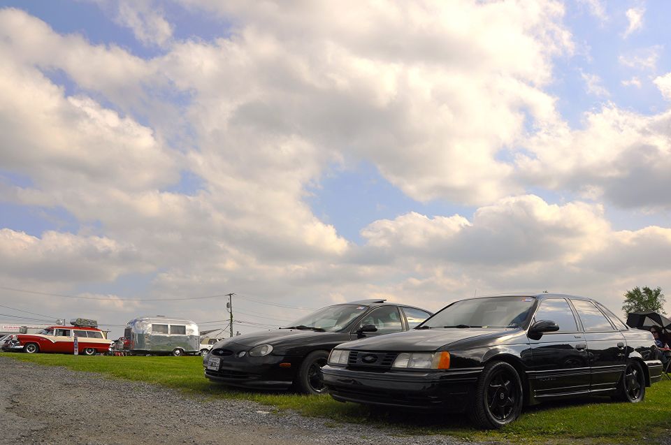 Black sedan parked on gravel, front 3/4 view. Visible text 'SUPERMOTORS'