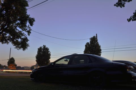 1997 Ford Taurus - A black sedan is parked on grass at dusk, with a distant sign indicating "Work In Progress".