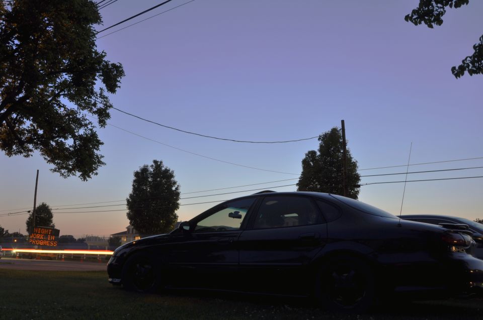 Black sedan parked on grass, side view, with a "Work In Progress" sign in the background