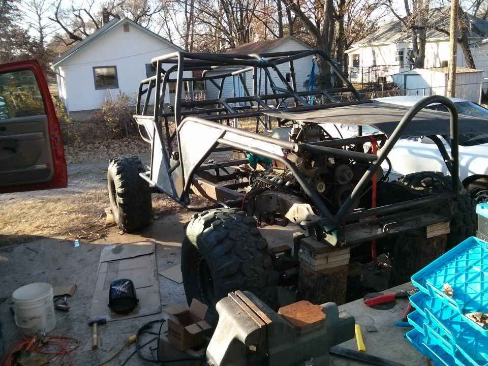 Custom off-road buggy frame with engine and large tires, partially assembled outdoors. Red door visible on left