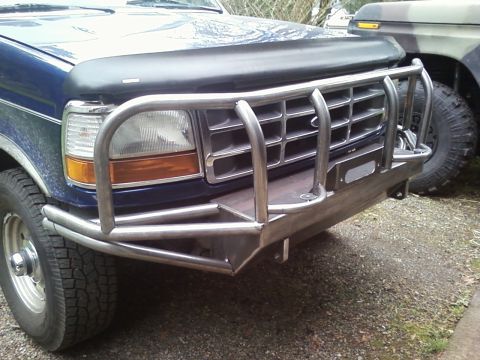 1989 Ford Bronco - A blue Ford truck is fitted with a heavy-duty brush guard and custom bumper, parked next to a camouflage vehicle.