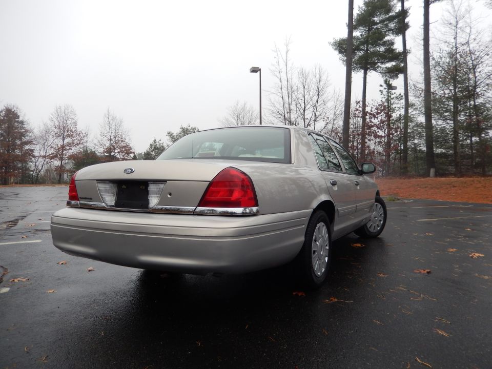 Rear view of a silver 2001 Ford Crown Victoria parked on a wet asphalt lot with trees in the background