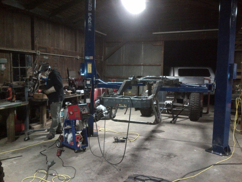 Jeep CJ frame on a lift with a welder working nearby and a silver pickup truck in the background
