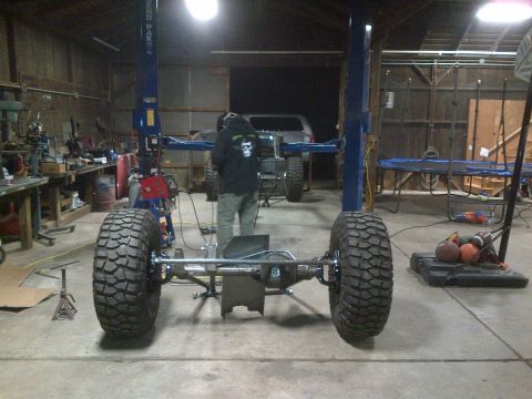 1985 Jeep CJ7 - The chassis of a Jeep CJ is elevated on a lift, its large off-road tires and suspension components.