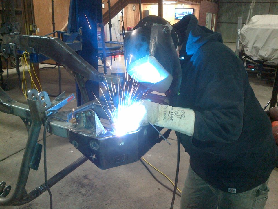 Jeep CJ frame section with "JEE" stamped on it, being welded by a person in a mask and gloves