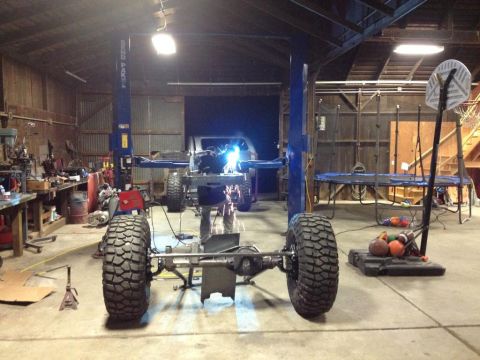 1985 Jeep CJ7 - A Jeep CJ frame is undergoing welding work on a lift in a workshop, with large off-road tires attached.