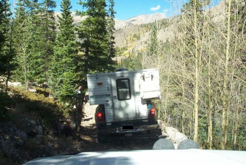 1990 Ford Bronco - Truck with camper shell navigates a rough mountain trail with trees and hills in the background.