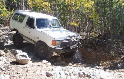 1990 Ford Bronco - This modified SUV navigates a challenging, rocky off-road path surrounded by trees.