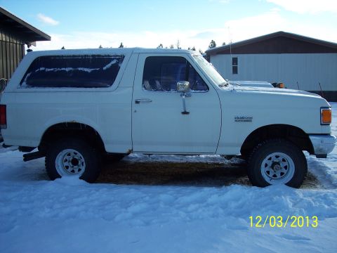 1990 Ford Bronco - A white Ford Bronco is parked in a snowy area, showing its side profile with a building in the background.