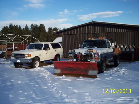 1973 Ford F-250 - Two trucks equipped with snow plows sit in the snow next to a barn on a sunny day.