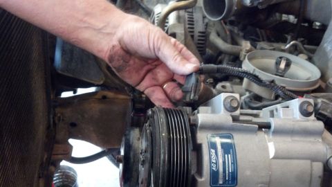 1993 Ford Bronco - Mechanic's hand disconnects a sensor in the engine compartment, near the AC compressor and radiator.