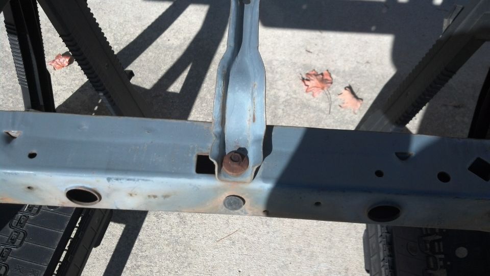 Gray metal vehicle frame with rust and bolt visible. Tire tread and shadows on concrete