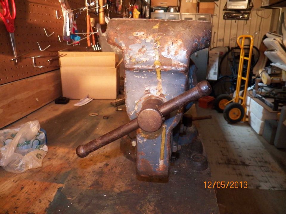 Rusty bench vise with a large handle, set on a wooden workbench. Tools hang on a pegboard behind it