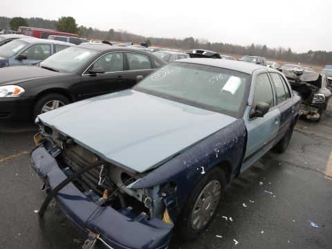 1991 Ford LTD Crown Victoria - A damaged blue and light blue sedan sits in a junkyard with other vehicles visible in the background.