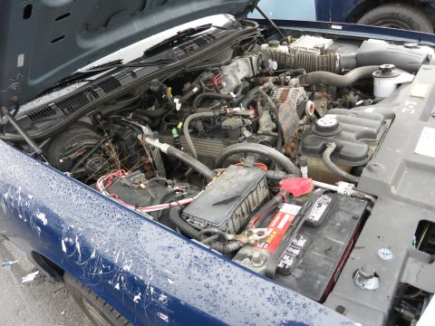 1991 Ford LTD Crown Victoria - The engine compartment of a blue vehicle, showing the battery and various engine parts. The car appears to be wet.
