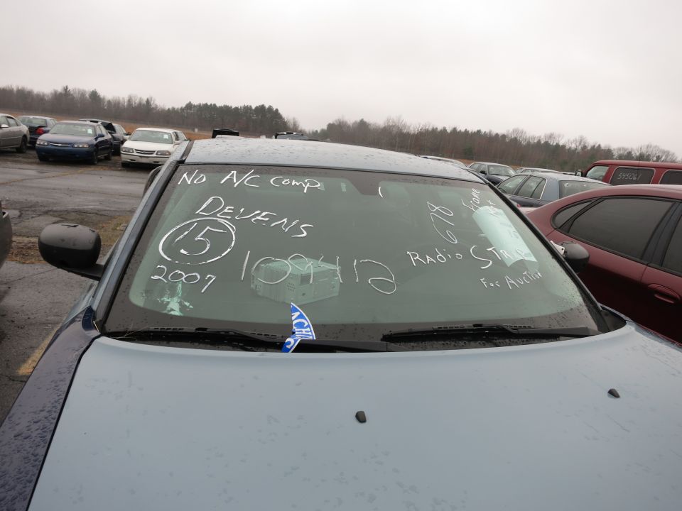 Light blue car windshield with white writing: 'NO A/C comp', 'DEVENS', '15', '2007', '109412', '828', 'Radio STR', 'For Auction'. Blue ACH tag