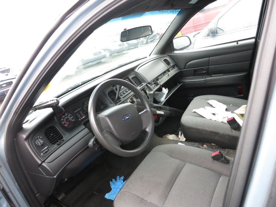 Interior of a Ford Crown Victoria showing the dashboard, steering wheel, and seats. The glove compartment is open and the interior appears messy