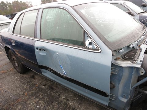 1991 Ford LTD Crown Victoria - Sedan in junkyard with mismatched paint and missing front end parts. Windshield has writing on it.