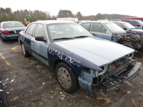 1991 Ford LTD Crown Victoria - A two-tone sedan sits in a junkyard, missing its front bumper and grille, with other vehicles visible in the background.