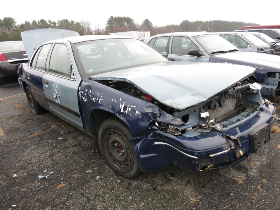 Damaged blue and light blue Ford Crown Victoria with hood open, front end crushed, parked in a junkyard