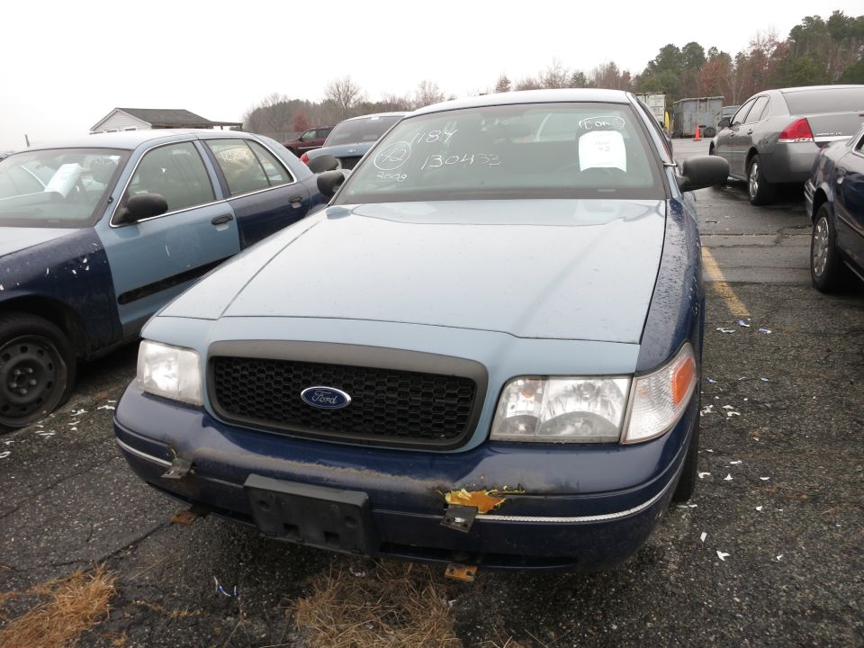 Front view of a blue and light blue Ford Crown Victoria with writing on the windshield. Another blue car is to its left