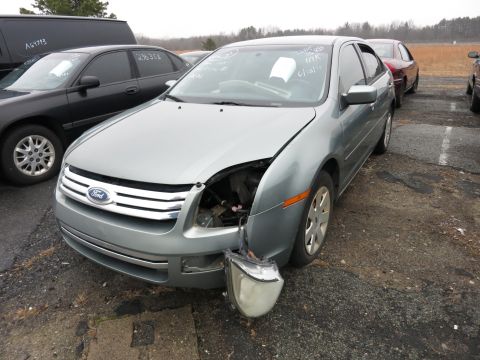 1991 Ford LTD Crown Victoria - A damaged silver Ford Fusion sits in a lot, missing its driver's side headlight and part of its grille.