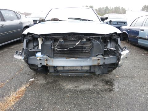 1991 Ford LTD Crown Victoria - The front of a damaged white car, missing its bumper and grille, is seen in a junkyard with other vehicles.