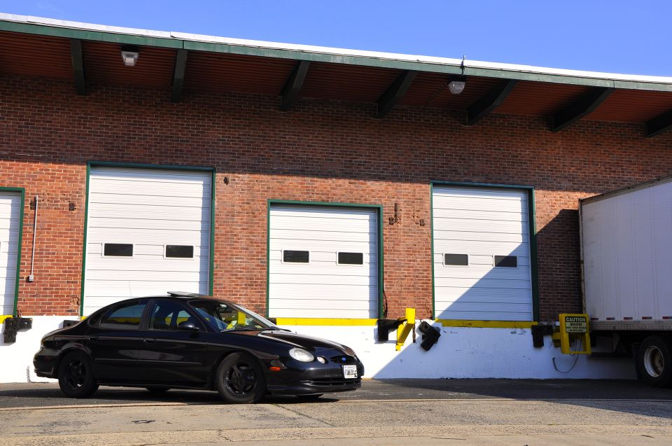 Black sedan parked in front of a brick building with three garage doors. A semi-truck is parked to the right