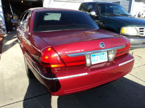 2004 Mercury Grand Marquis - Rear view of a red 2004 Mercury Grand Marquis GS sedan parked in front of a garage, with a green pickup truck visible behind it.