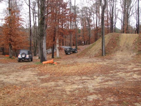 1979 Ford Bronco - Two off-road vehicles are in a wooded area with a dirt ramp, during autumn.
