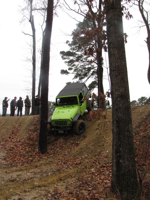 1979 Ford Bronco - A group of people watch as a green Jeep Wrangler navigates a steep, muddy incline in a wooded area.