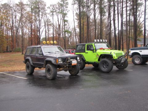 1979 Ford Bronco - Two modified SUVs, a dark-colored Jeep Cherokee and a lime green Jeep Wrangler, are parked on a wet lot with trees in the background.
